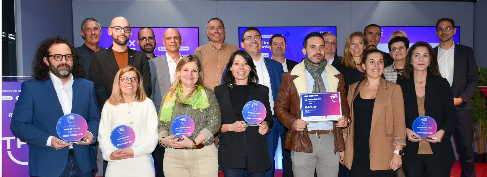 image présente les gagnants du Prix TPE, réunis lors de la cérémonie officielle. Ils posent fièrement avec leurs distinctions : des trophées circulaires aux couleurs vives (bleu, violet, rose) et un panneau rectangulaire indiquant le nom d’une entreprise primée. En arrière-plan, on distingue un décor moderne avec des écrans lumineux affichant le logo et les couleurs de l’événement, dominées par des tons violets et bleus. L’ambiance est professionnelle et festive, reflétant la reconnaissance des talents et des réussites entrepreneuriales.